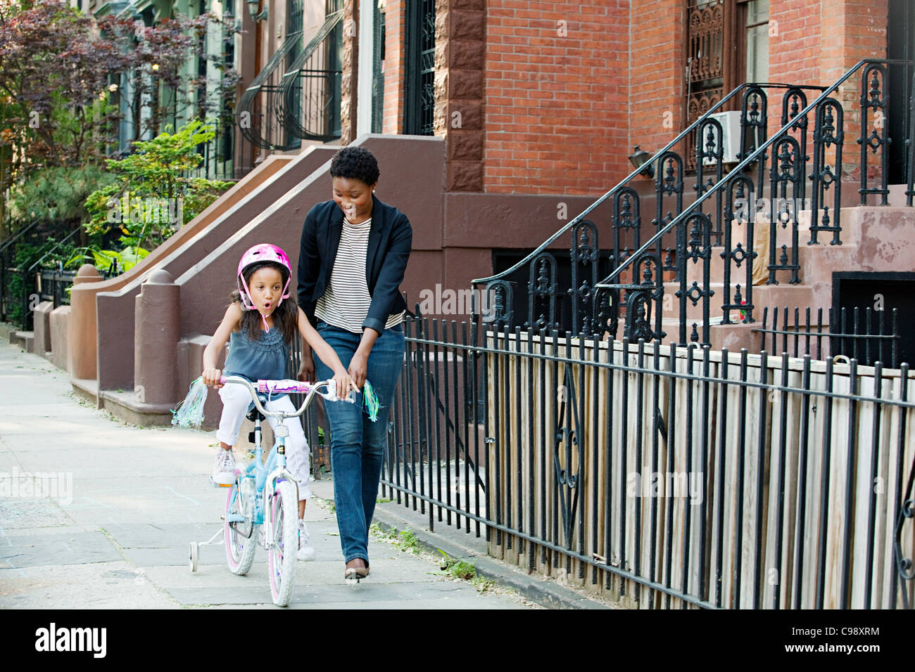 Daughter learning to ride bicycle along sidewalk with mother Stock Photo