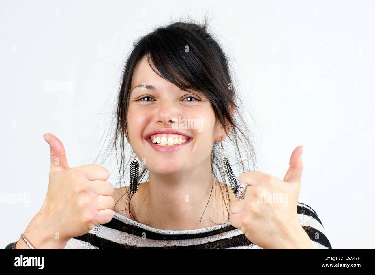 Funny candid shot of a young dark haired woman smiling and making the two thumbs up sign of approval, studio shot over white. Stock Photo