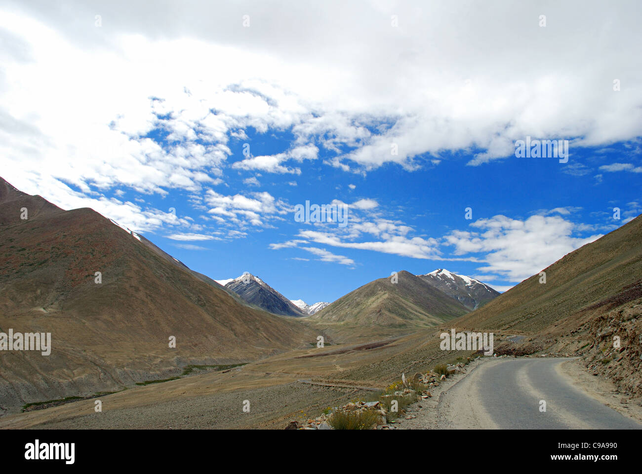 The road to Leh, on the right hand bottom corner. Snow clad Himalayan Mountain Ranges, White clouds and Blue sky in the backgrou Stock Photo