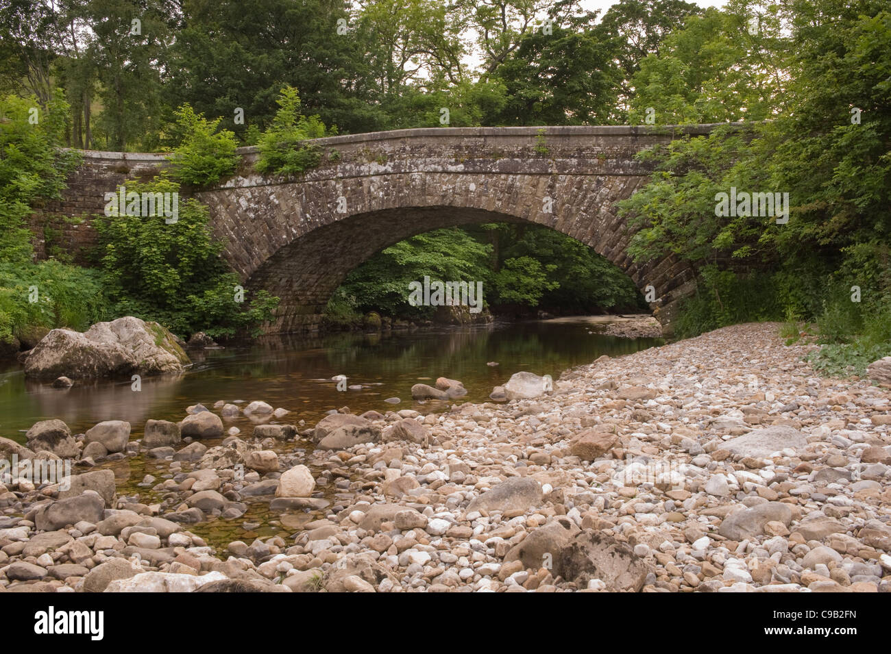 Old stone road bridge (1 arch) spanning shallow flowing water of River Wharfe (quiet scenic wooded valley) - Hubberholme, Yorkshire Dales, England, UK Stock Photo
