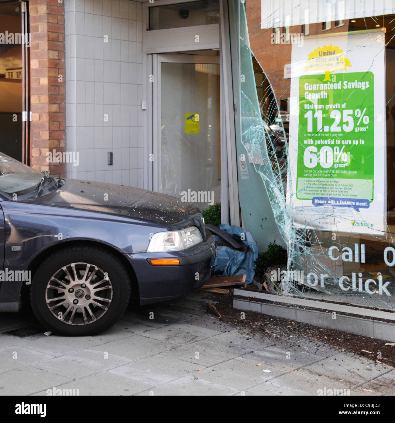 Road traffic accident crash car mounts pavement in shopping street scene & hits window of building society shop front smashed broken glass England  UK Stock Photo