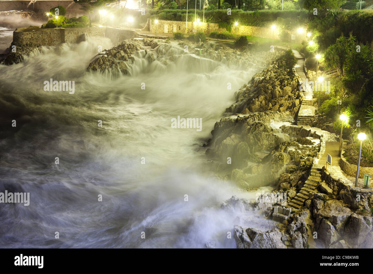 Night time exposure of a rough sea crashing down on a rocky shoreline alongside a lighted footpath. Nice, French Riviera, France. Stock Photo
