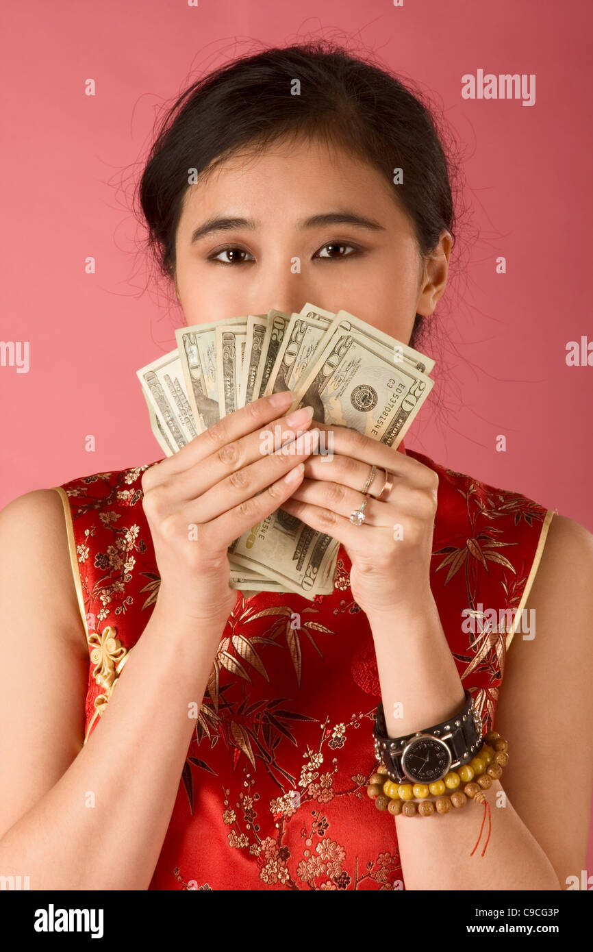 Portrait of young Chinese woman in traditional clothes (cheongsam) holding US twenty dollars bills, covering her face by them. Stock Photo