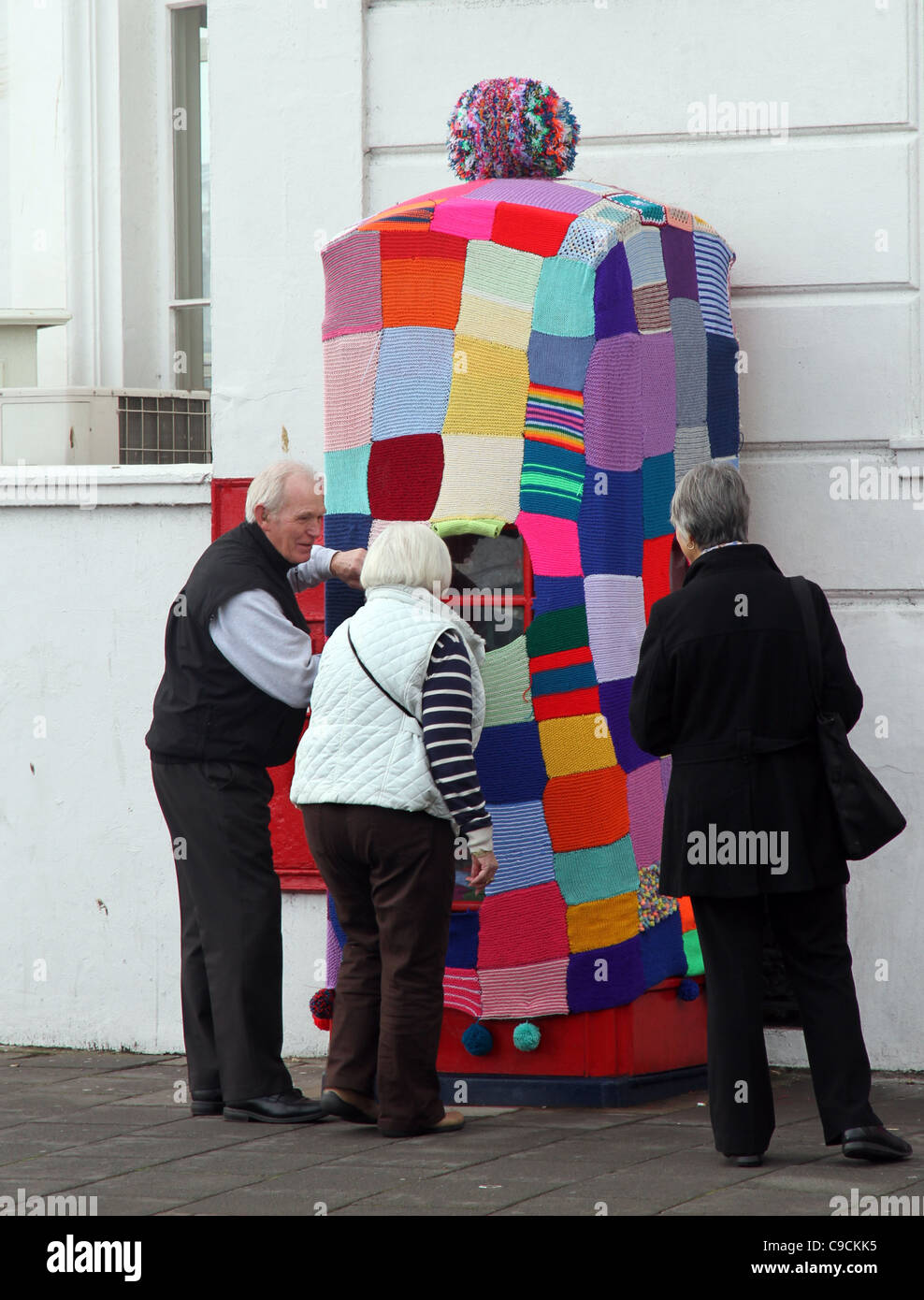 Three people looking at a red public telephone kiosk that had been covered over with knitting like a big tea cosy. Stock Photo