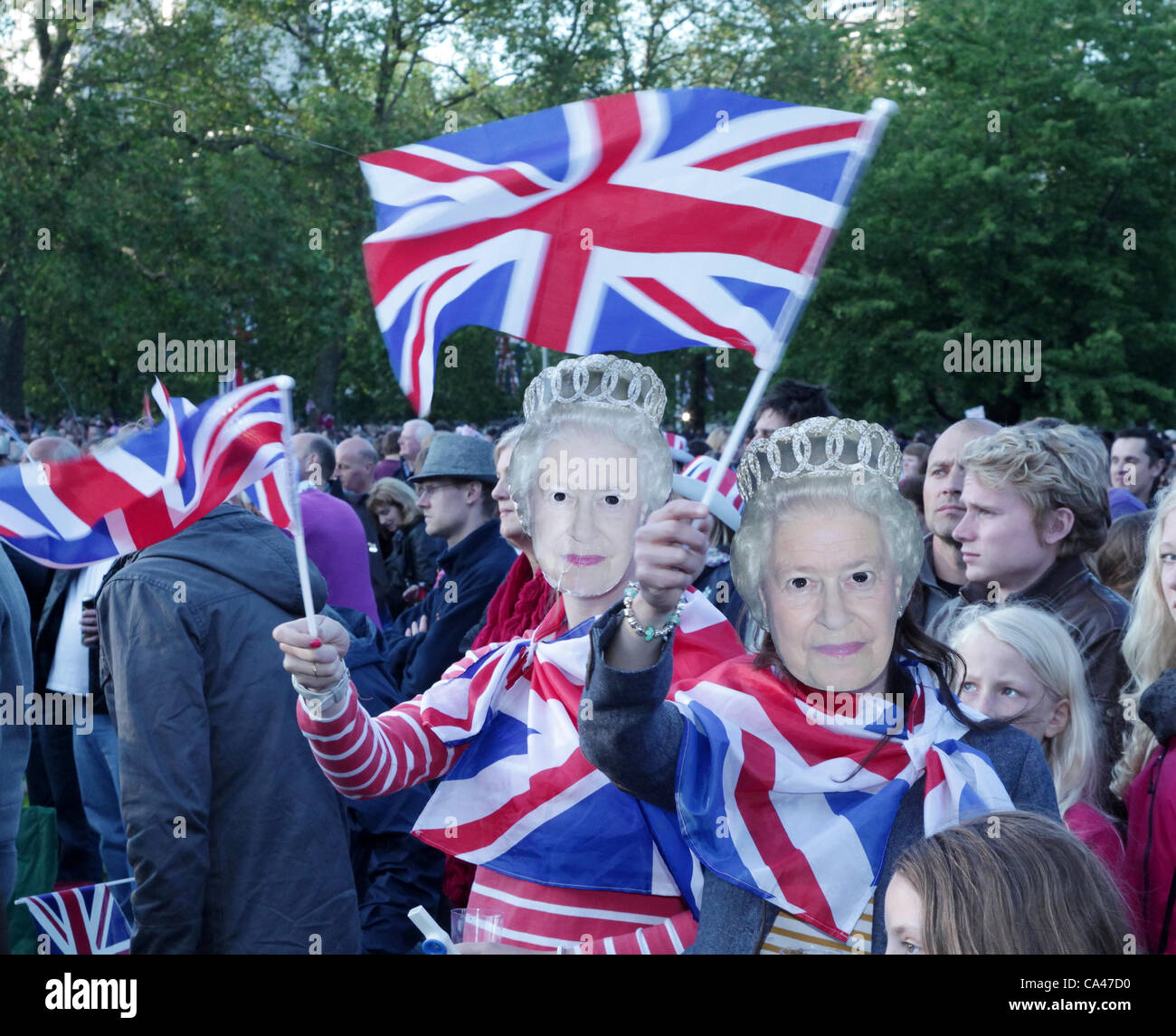 London, UK. June 4, 2012. Two teenager girls with 'Queen' masks enjoying the music at the Concert to celebrate The Queen's Diamond Jubilee in St. James's Park watching the big screens, waving flags. Stock Photo