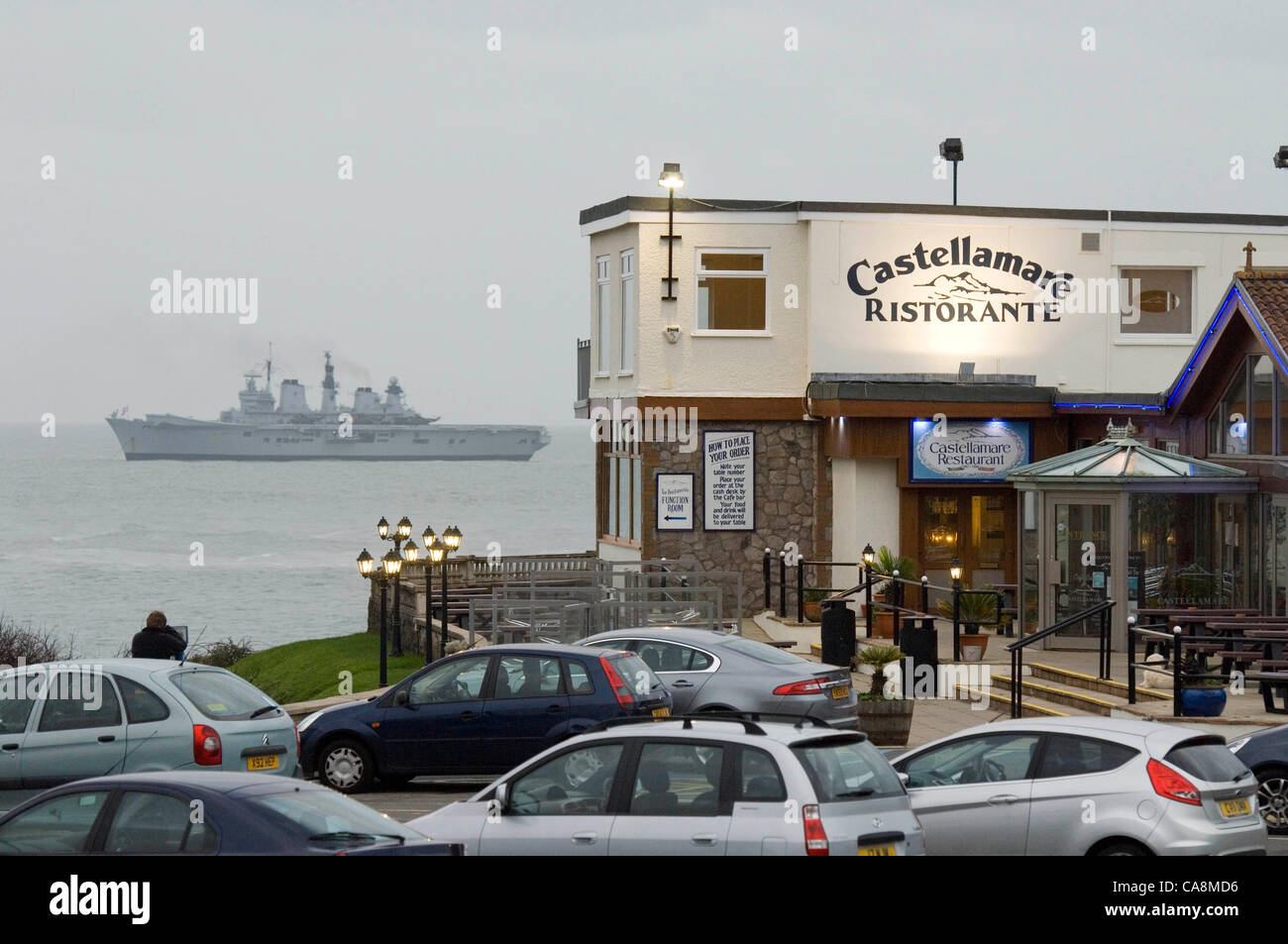 The Invincible-class light aircraft carrier -  Royal Navy - HMS Illustrious lays anchor off the coast of Swansea on 3rd December 2011 with Castellamare restaurant in Bracelet Bay in the foreground. Stock Photo