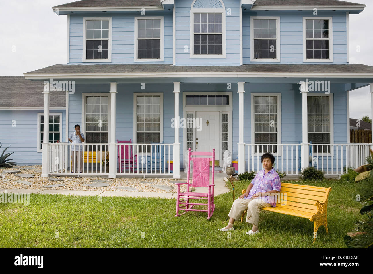 Mature Asian woman sitting on front lawn of house with husband smoking on porch Stock Photo