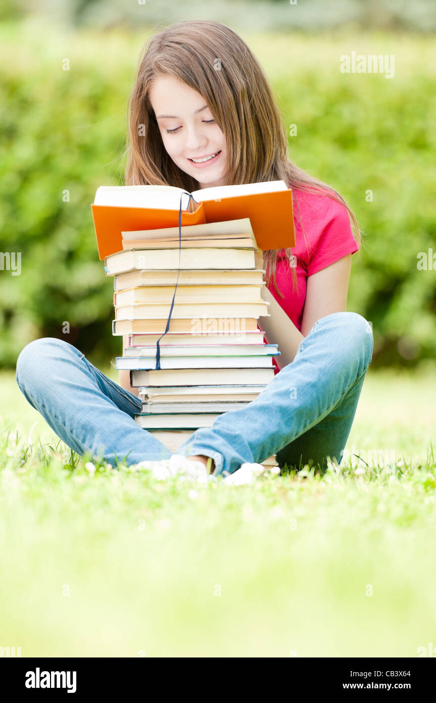 beautiful and happy young student girl sitting on green grass, pile of ...