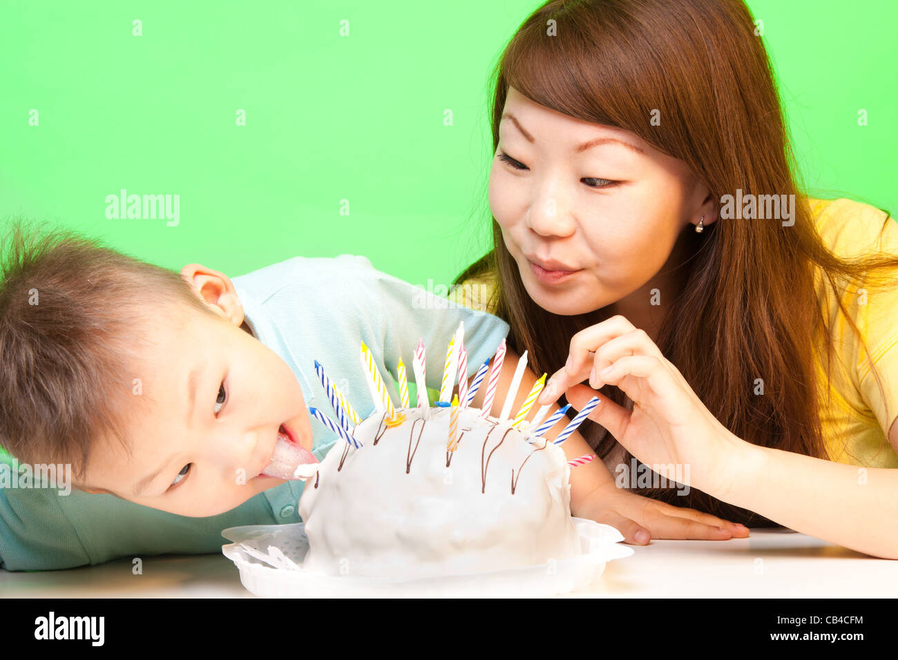 Boy licks his birthday cake with mother nearby and a lot of candles in it Stock Photo