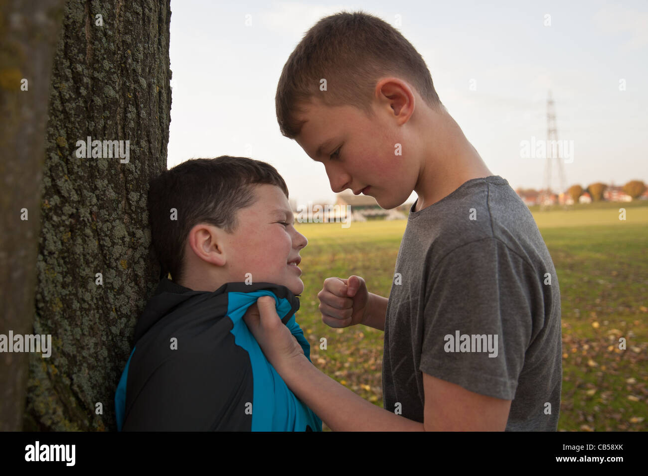 An older boy bullying a younger boy with his fist clinched holding him up against a tree. Stock Photo