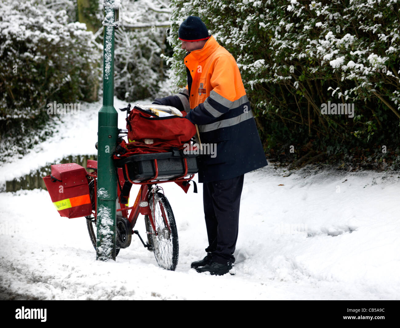 Postman Sorting Out Post Bag With Bicycle Leaning Against Lamp Post In The Snow Surrey England Stock Photo