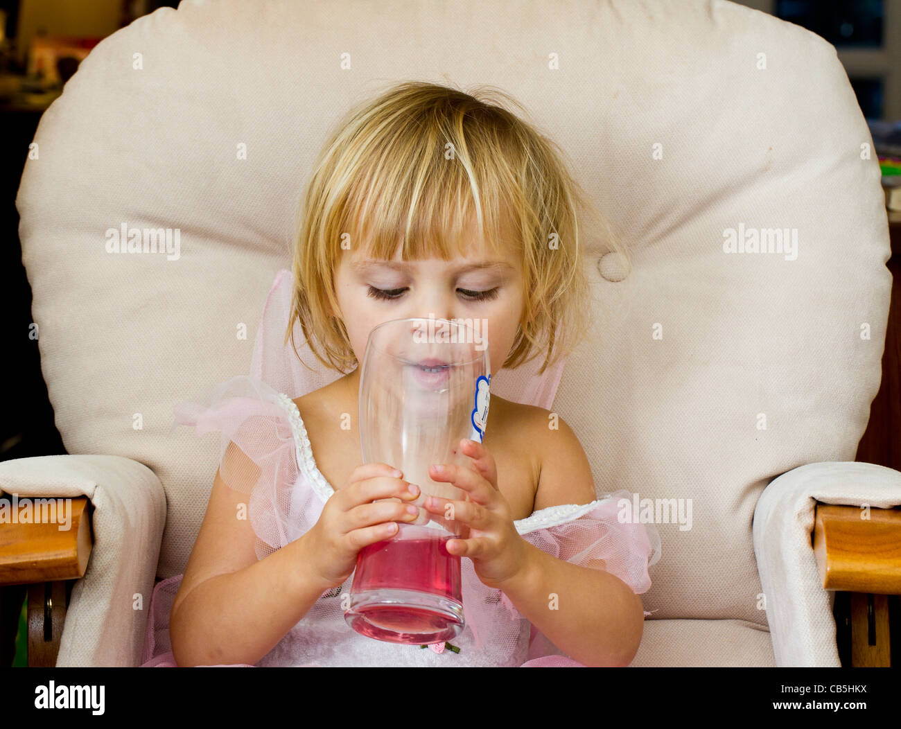 Little girl seated in a big chair, drinking a pink (blackcurrant) drink from a pint glass Stock Photo