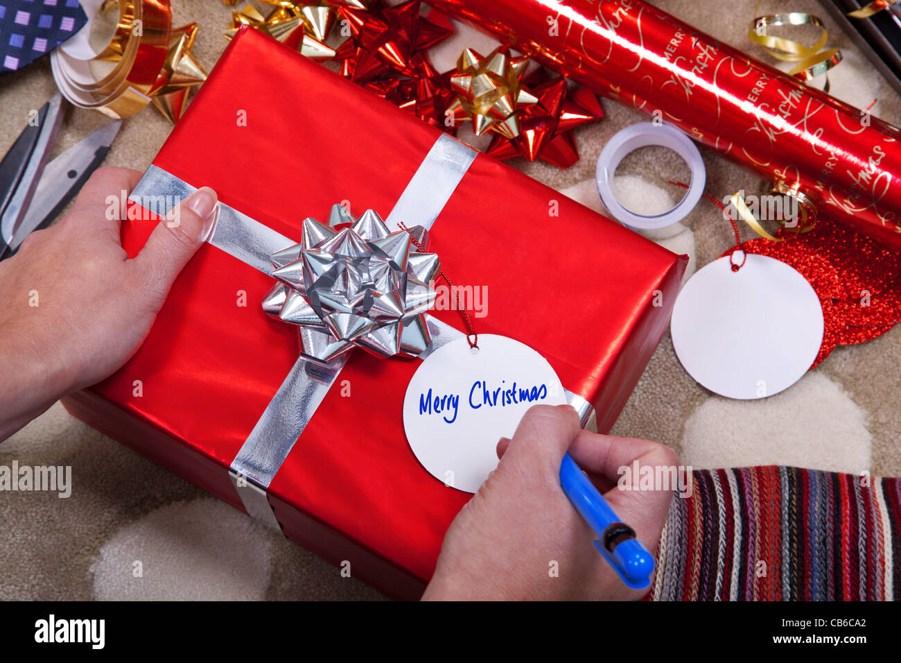 Photo of a woman writing Merry Christmas on a gift tag on a red present with silver ribbon and bow. Stock Photo