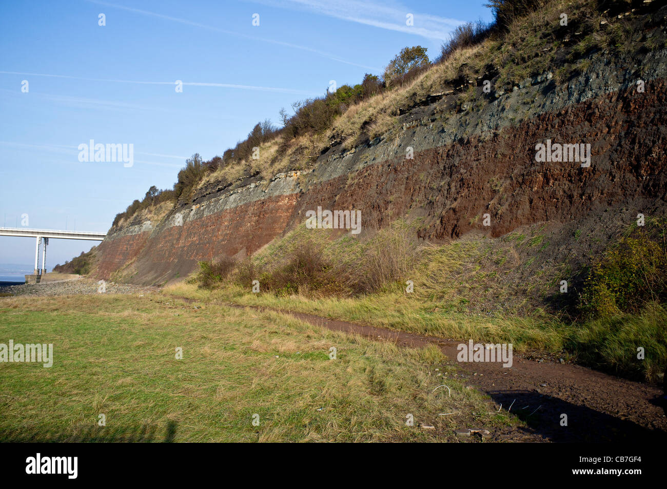 Red and green mudstone, shales and limestone at the SSSI site Aust Cliff, with the old Severn bridge in the background. Stock Photo