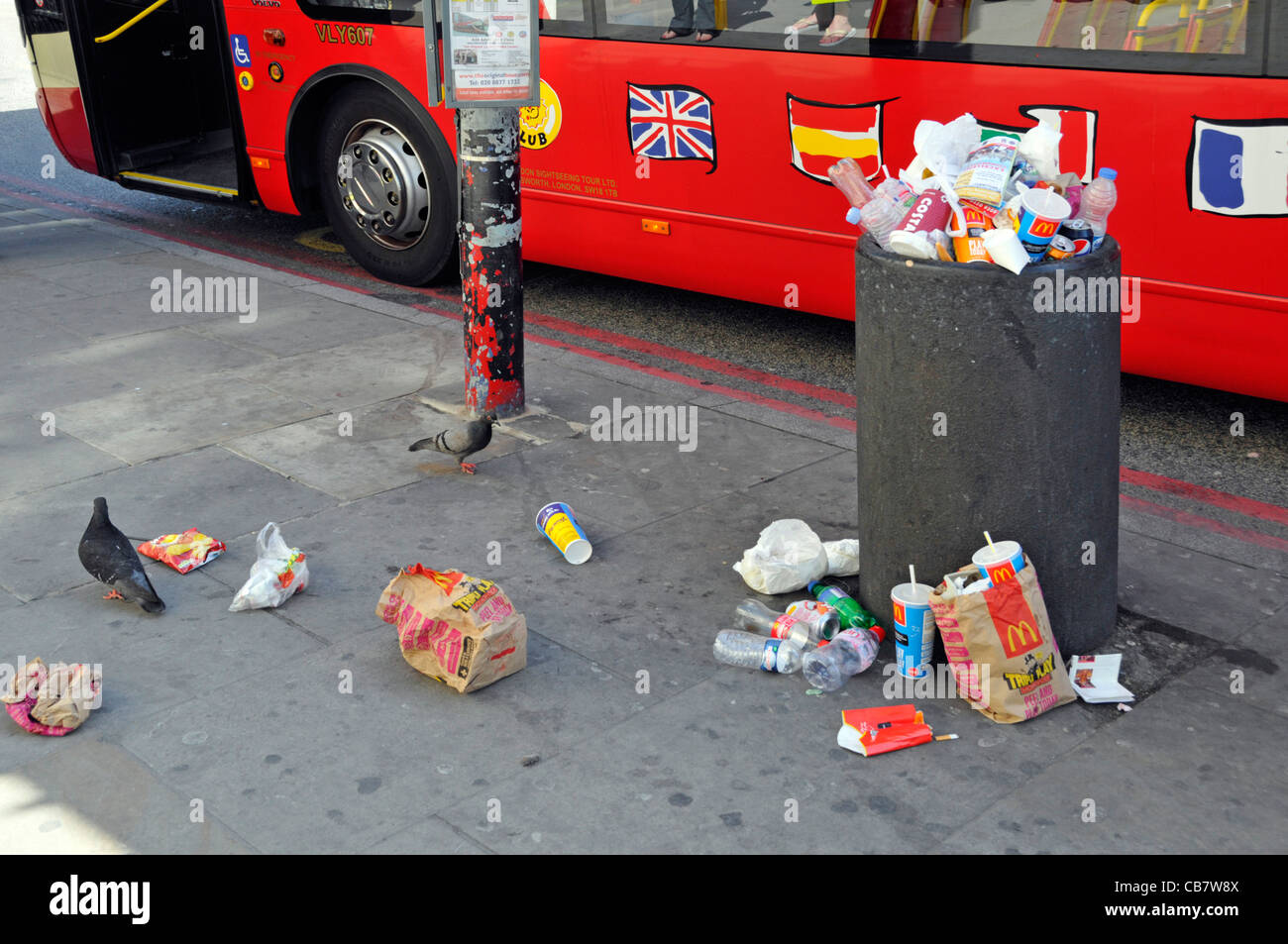 Waste management required to littering scene at street refuse bin full of rubbish garbage litter & trash spilling on pavement beside London tour bus Stock Photo