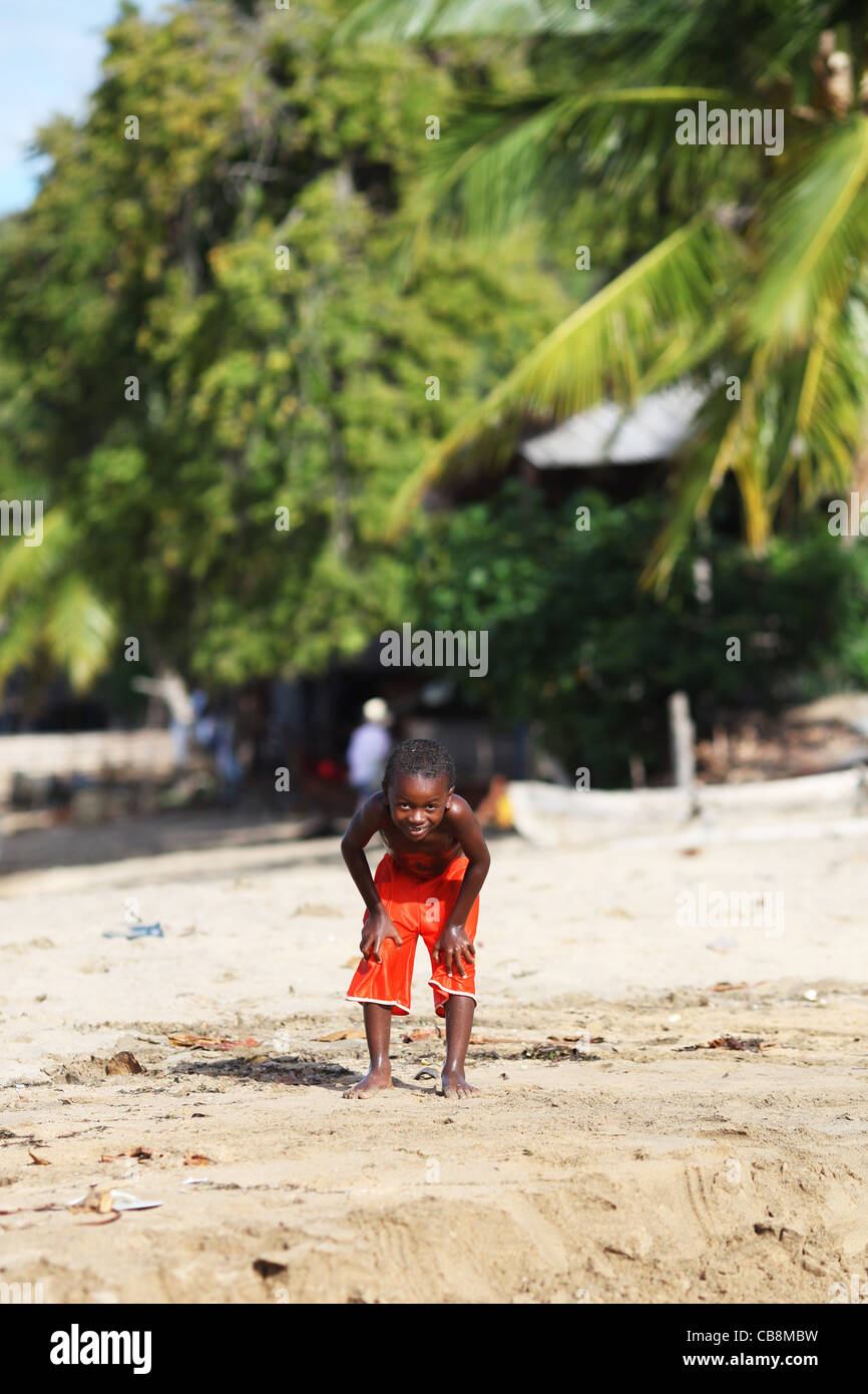 A young boy with in hands on his knees, in orange shorts or trunks, smiling at the beach, Ampangorinana, Nosy Komba, Madagascar Stock Photo