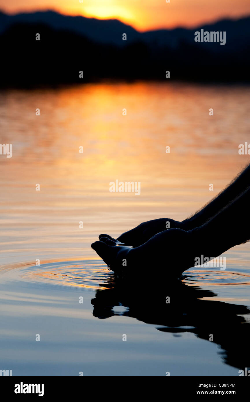 Cupped hands scooping up water in a still lake at sunrise in India. Silhouette Stock Photo