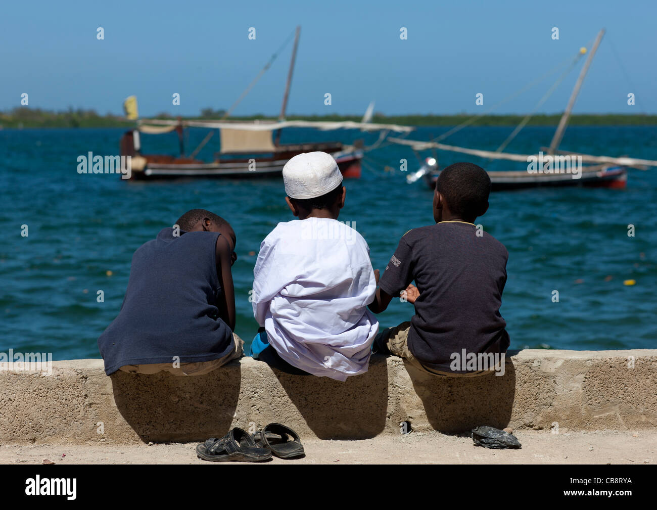 Three Little Muslim Boys Chatting Alongside The Dockside, Dhows In Background, Lamu, Kenya Stock Photo