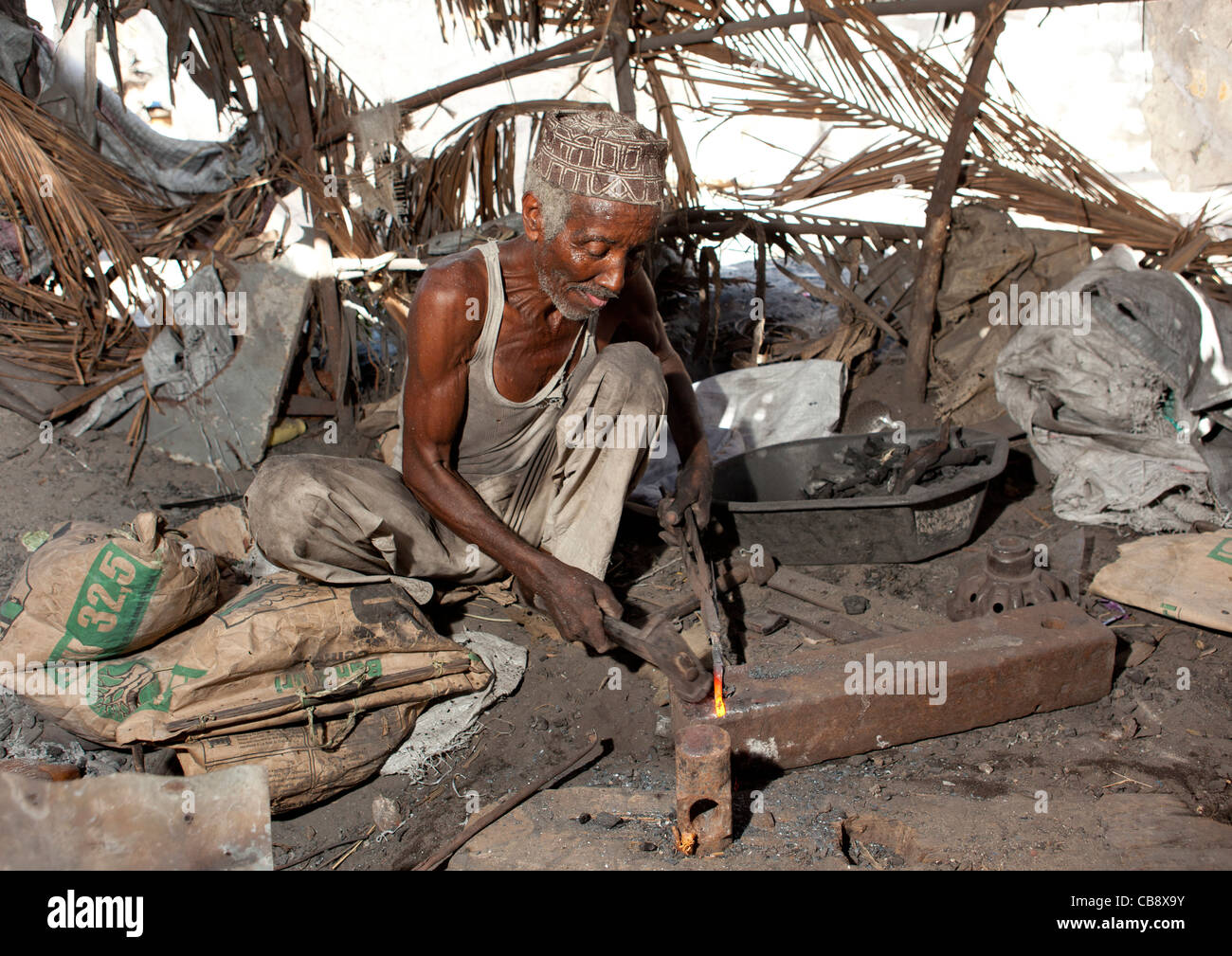 Blacksmith Sitting On The Floor With Kofia Sweating While He Forges Nail, Lamu, Kenya Stock Photo