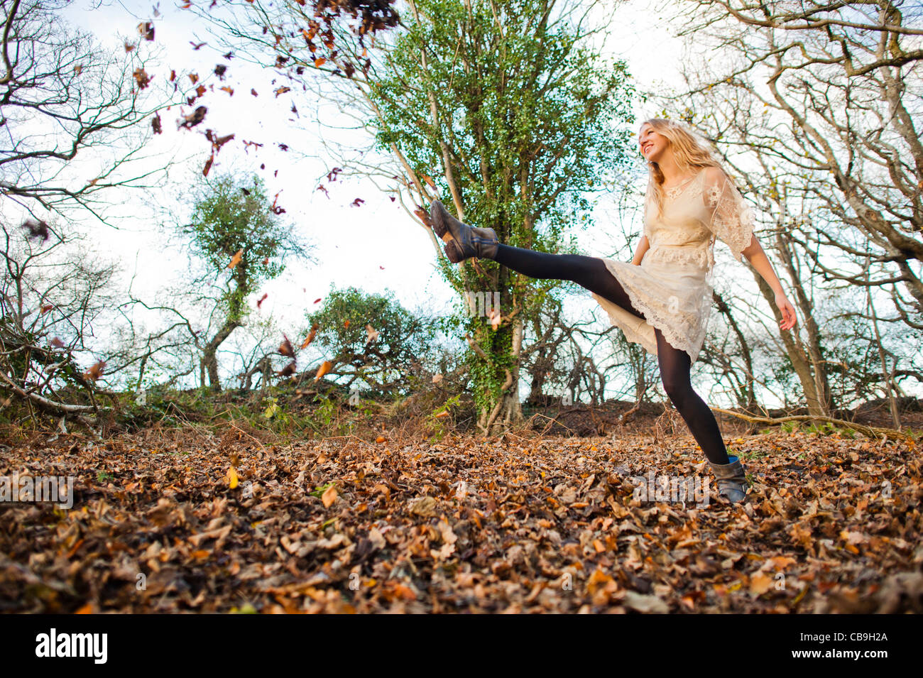 a slim blonde woman girl alone in woodland autumn afternoon daytime kicking up dry leaves, UK Stock Photo