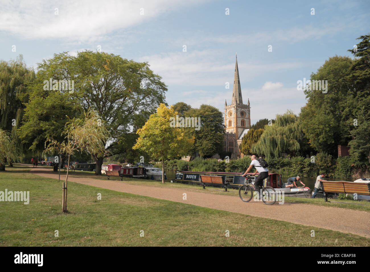 Cyclist passing canal boats moored on the River Avon with Holy Trinity Church behind, Stratford Upon Avon, Warwickshire, UK. Stock Photo