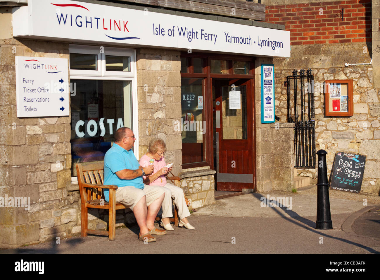 Mature couple sat on bench enjoying ice creams outside Wightlink ferry terminal at Yarmouth, Isle of Wight, Hampshire UK in September Stock Photo