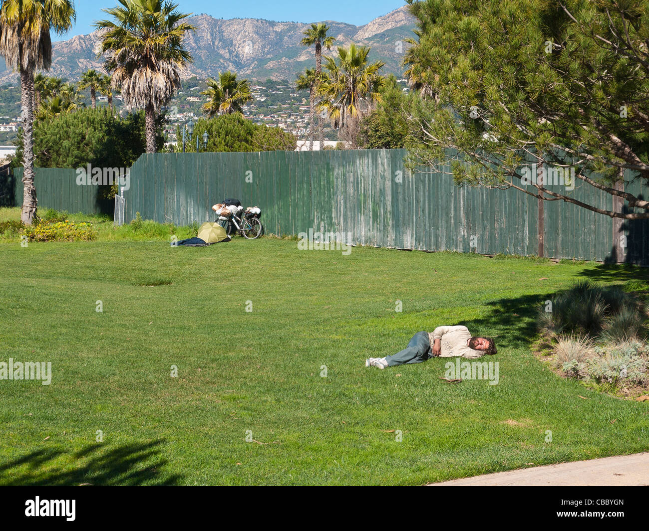 A thirty something homeless man sleeps midday on the green grass of a city park with his bicycle. Stock Photo