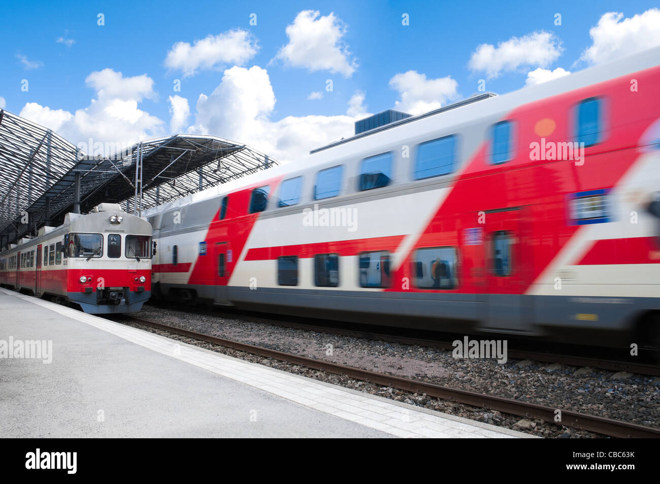 two trains in white and red color: one new, fast moving, and one old model at train station win blue cloudy sky in background Stock Photo