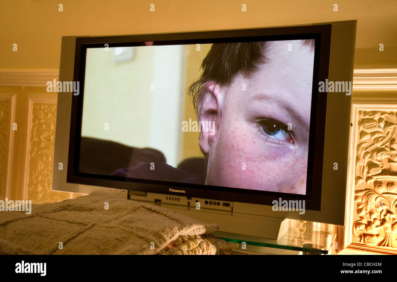 The right eye of a young man peers out from a flat screen television in a domestic lounge. Stock Photo