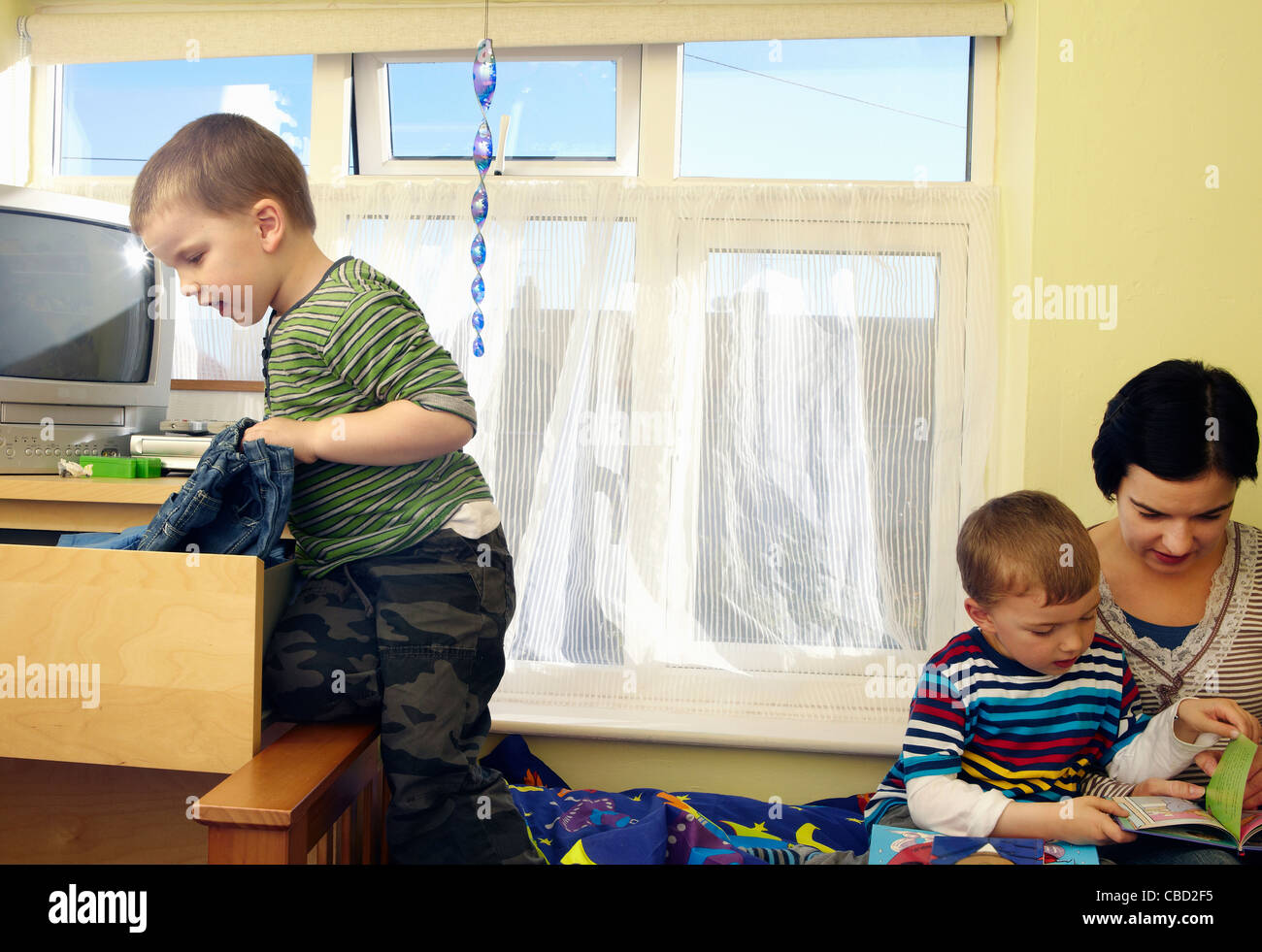 boy gets clothes out of drawer while mum reads to his brother Stock Photo