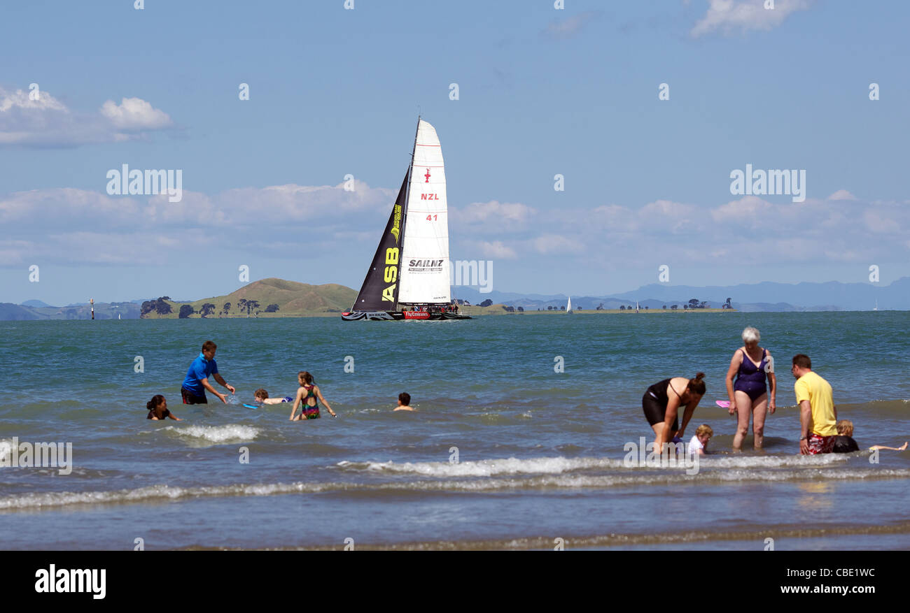 NZL 41 America's Cup Yacht passes families playing in the water at Cheltenham Beach, Auckland, New Zealand Stock Photo
