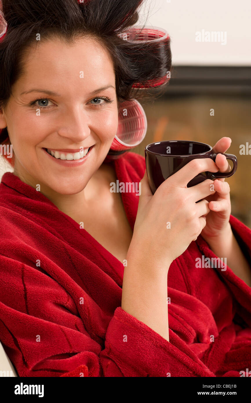 Woman with curlers holding mug sitting by fireplace red bathrobe Stock Photo