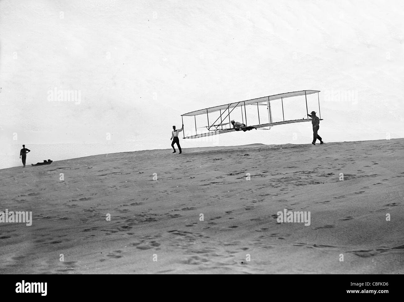 Wilbur in motion at left holding one end of glider, Orville lying prone in machine, and Dan Tate at right; Kitty Hawk Stock Photo