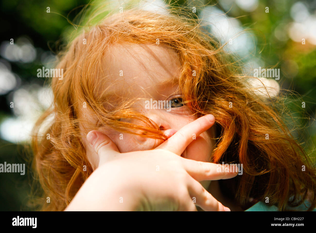 Little girl, aged 5, with her hand covering her mouth in a garden. Summer. Stock Photo