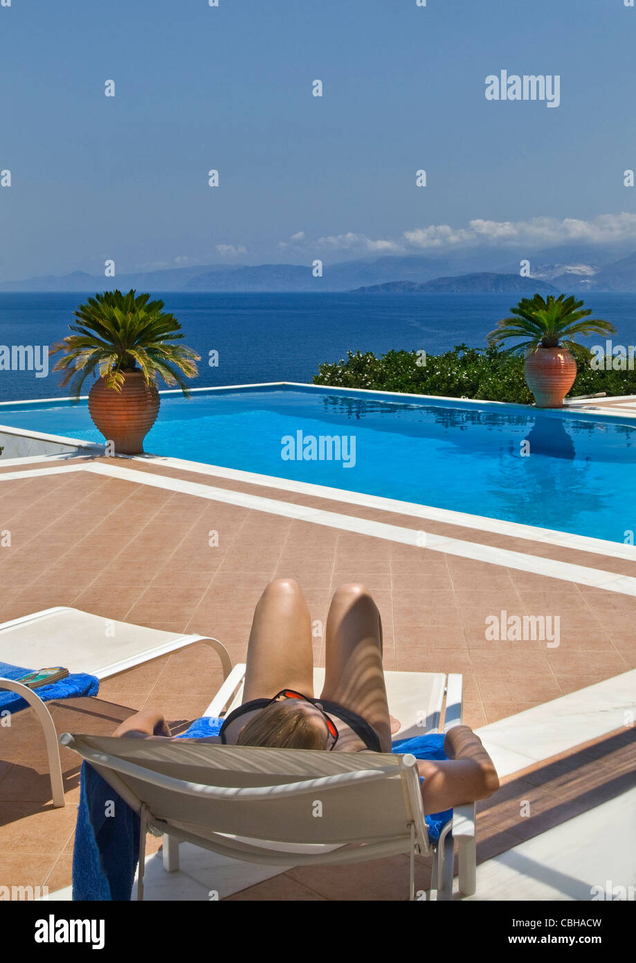 Holiday Woman infinity pool relaxing on vacation sun lounger with luxury infinity pool and coastal sea view behind Stock Photo