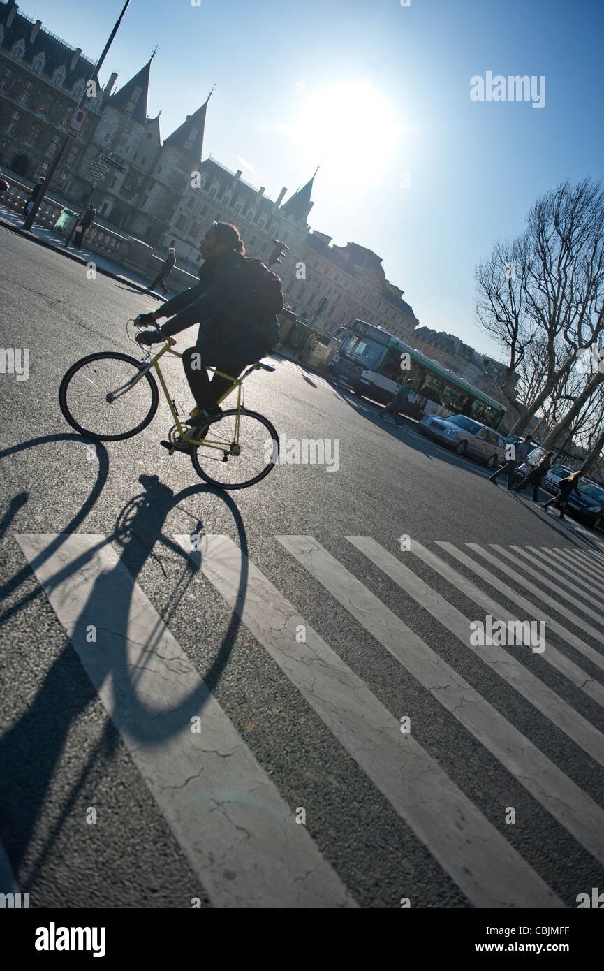 A man rides a bicycle on a main road in Paris, France, while the strong sun behind him casts a long shadow. Stock Photo