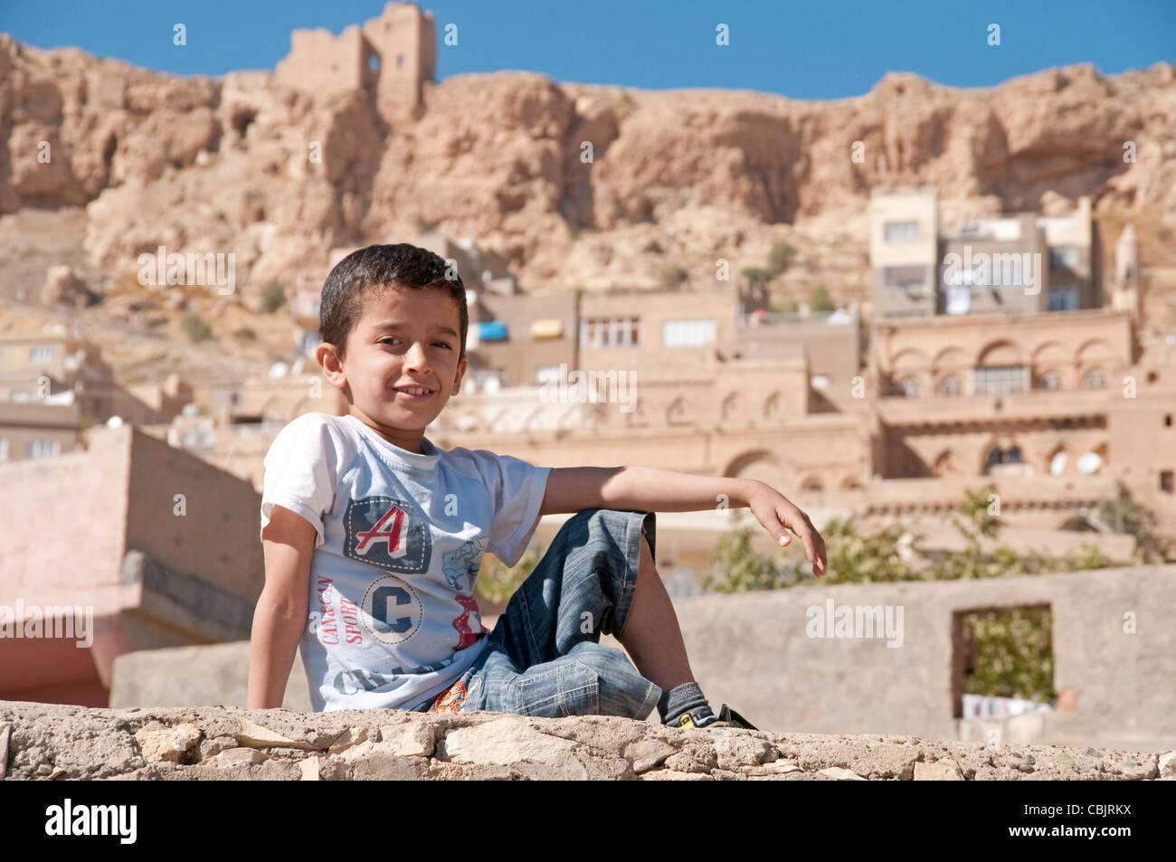 A young Kurdish boy sitting on a stone wall in view of the old city of Mardin in the eastern Anatolia region, southeastern Turkey. Stock Photo