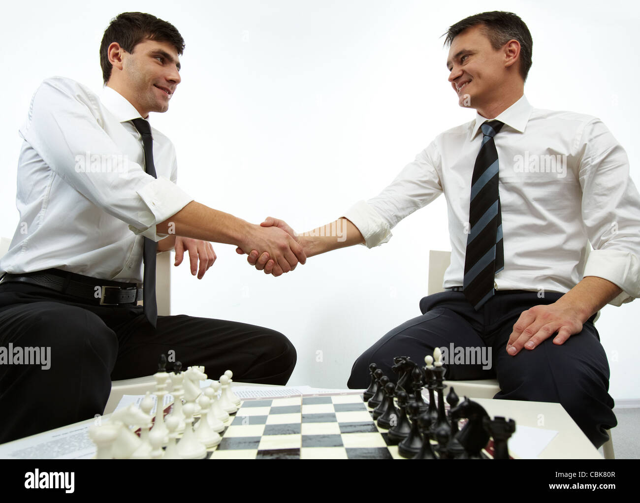 Two men handshaking with chess figures on chess board near by Stock Photo