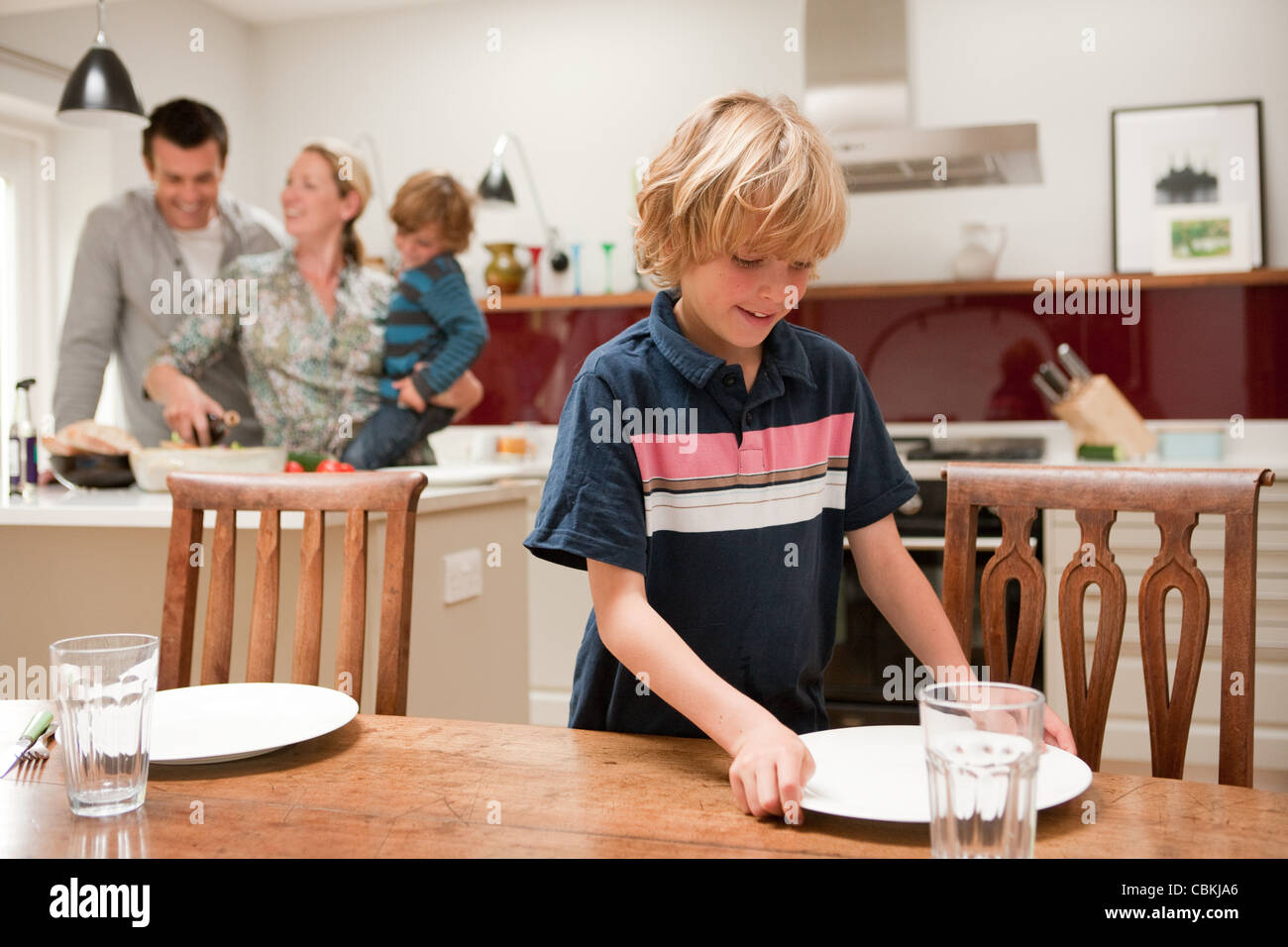 Son helping to lay table with parents and brother visible behind in kitchen Stock Photo