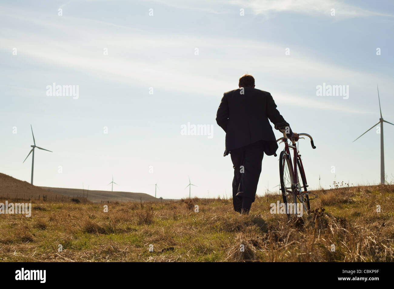 Man pulling bicycle uphill towards windfarm Stock Photo