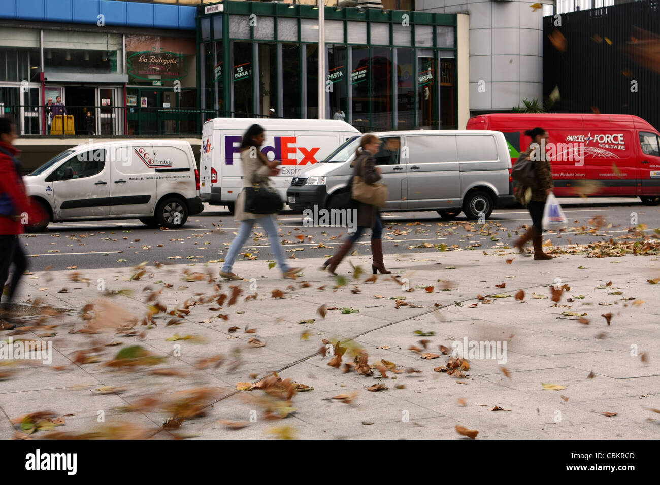 people caught in a gust of wind blowing leaves along a pavement and traffic in the background Stock Photo