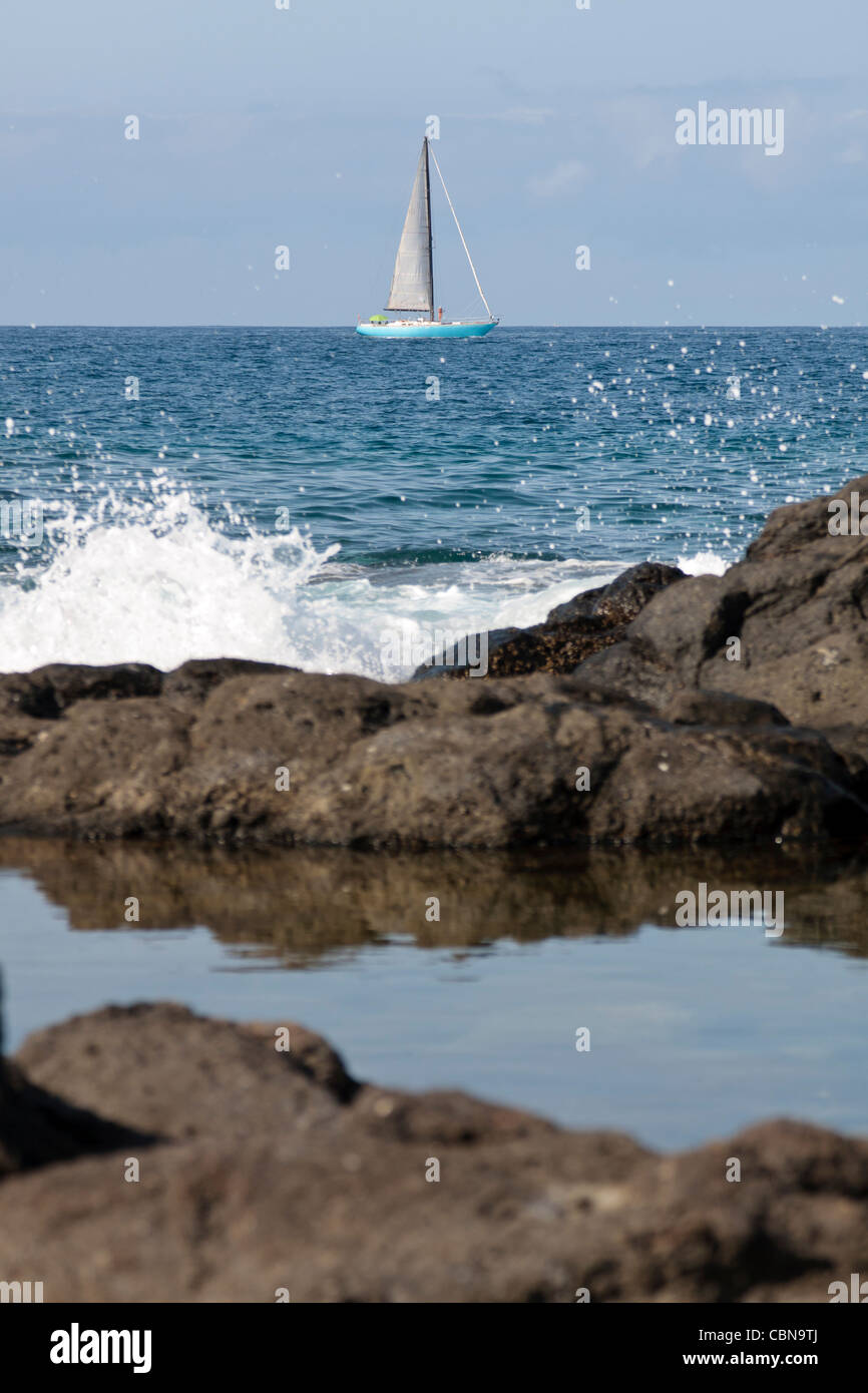 A yacht sails close to the coast as waves splash over the rocks in Playa San Juan Tenerife Canary Islands Spain Stock Photo