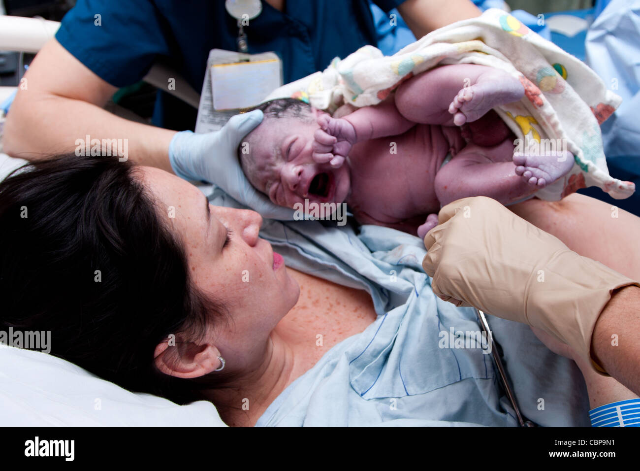 Mother in labor giving birth to baby new life in hospital. Infant covered in grease and lanugo is crying and held by nurses. Stock Photo