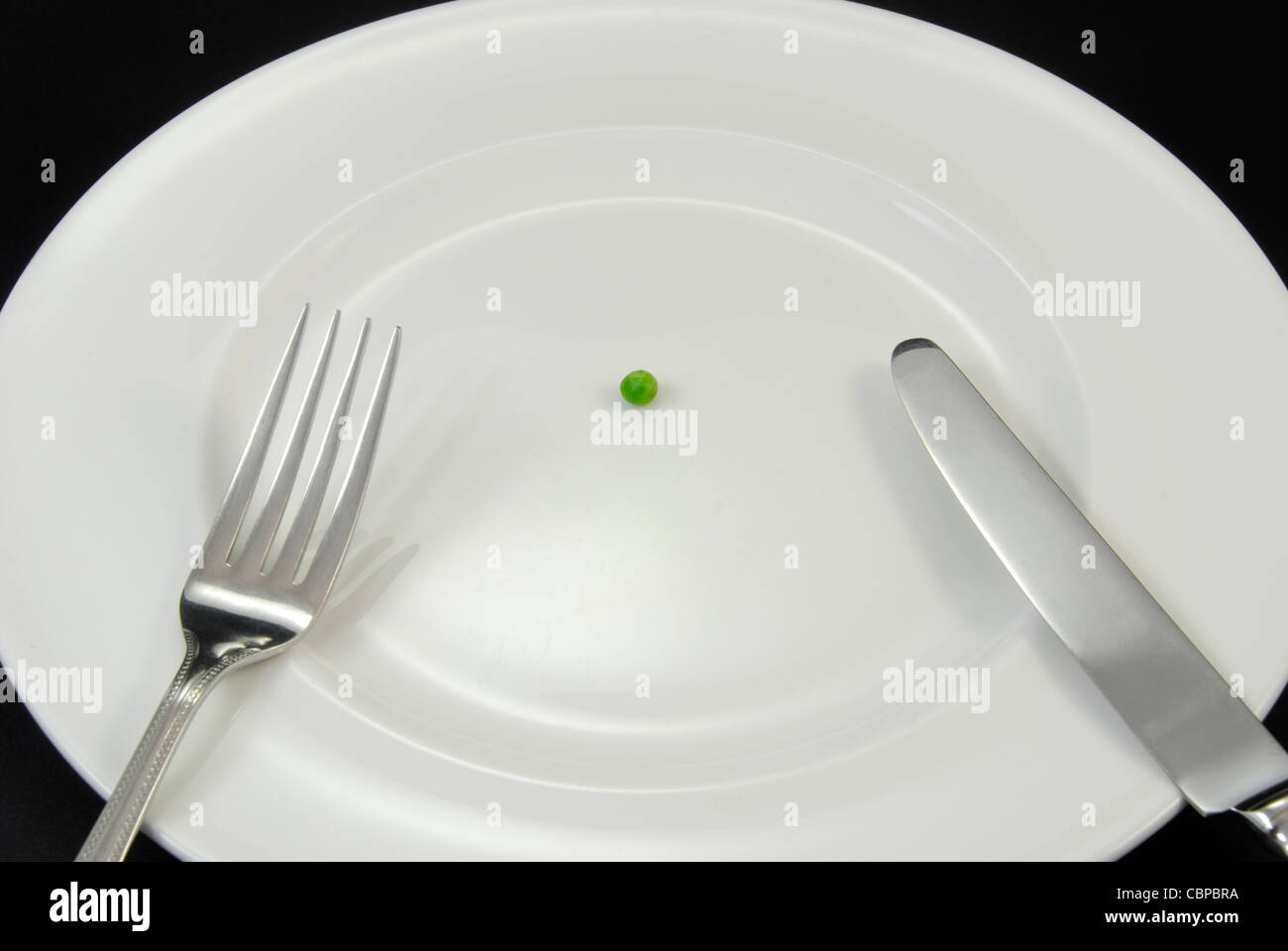 An exaggerated diet food menu consisting of a single pea on a plate with a knife and fork. Stock Photo