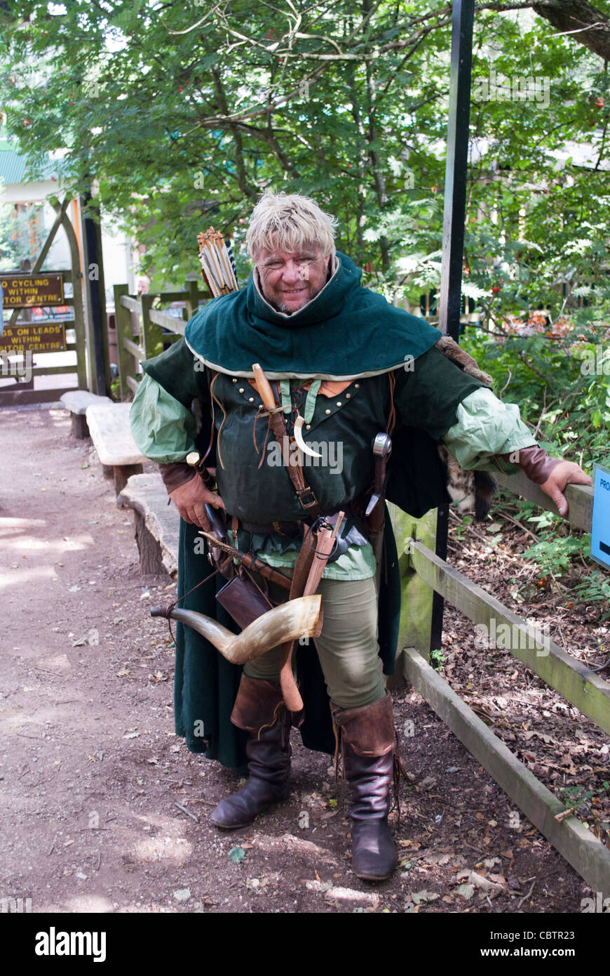 Large man dressed as one of Robin Hood's merry men with bow and arrows and hunting horn leaning on fence with trees behind Stock Photo