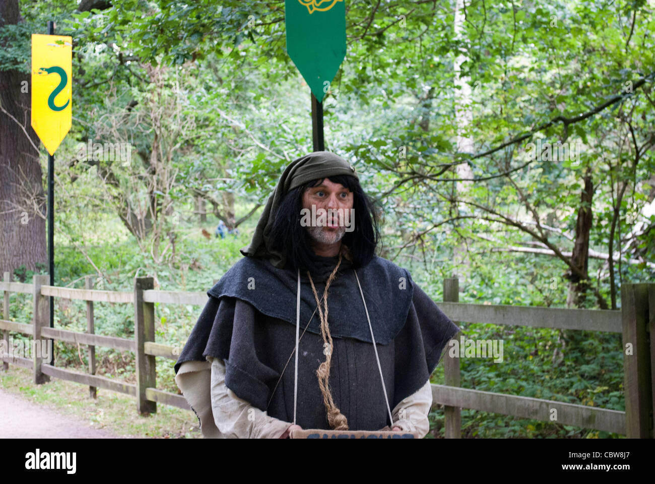 Man dressed as medieval peasant pulling a funny face walking past wooden fence with trees in background beneath green pennant Stock Photo