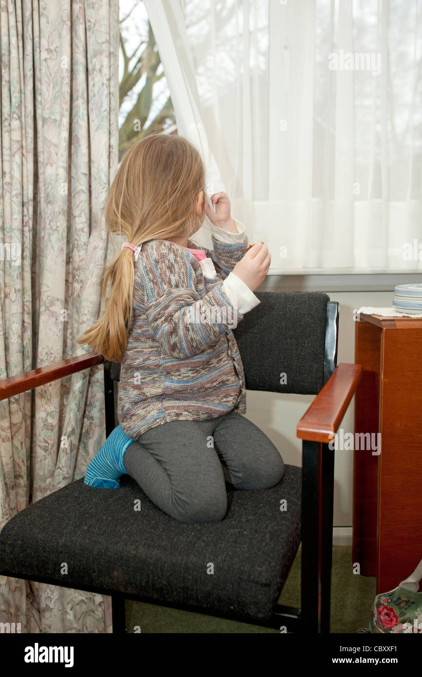 female toddler sat on chair looking out of window holding curtain Stock Photo