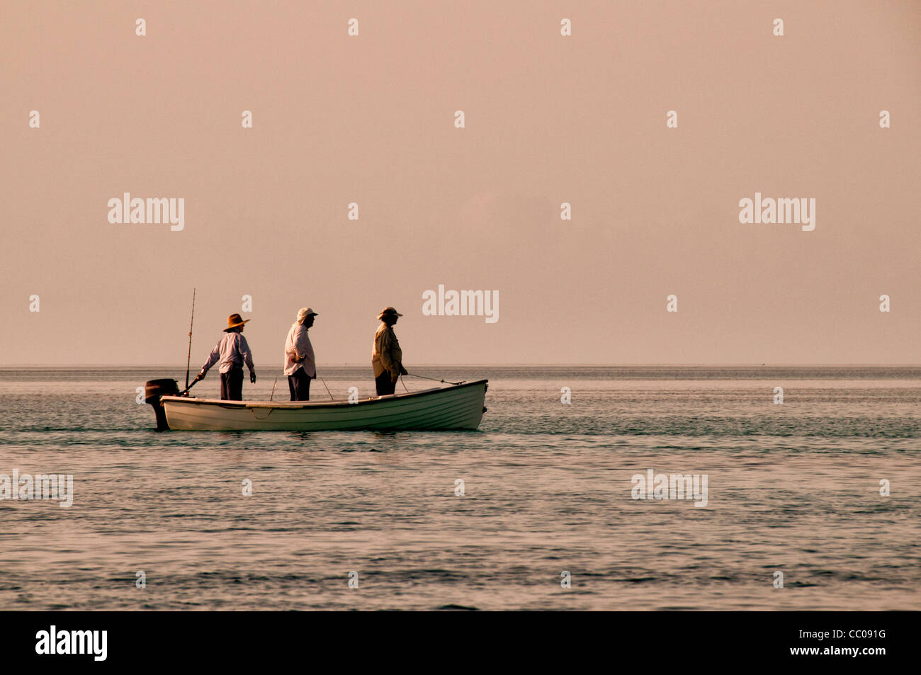 Three fishermen standing a small boat as they motor to a fishing spot on the edge of the reef at Swains Reef on the southern end of Australia's Great Barrier Reef. The later afternoon sun creates a warm, golden glow. Stock Photo
