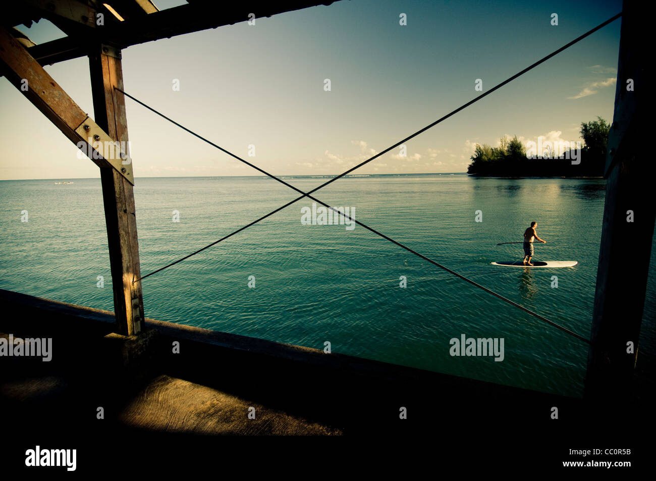 Paddle boarder on water paddling Hanalei Bay with view through pier