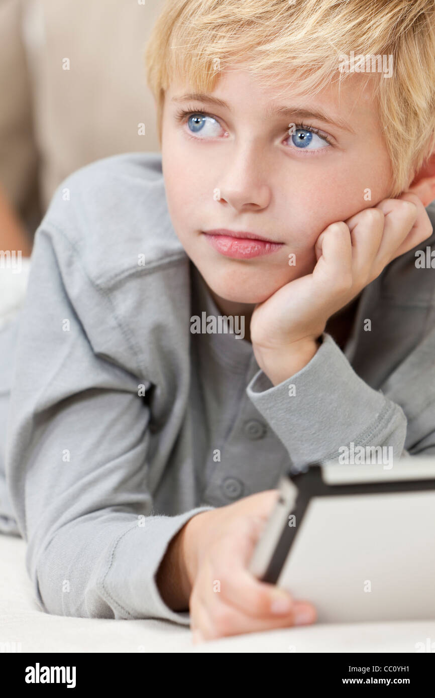 A young blond haired blue eyed boy child resting on his hands laying down on a sofa using tablet computer Stock Photo